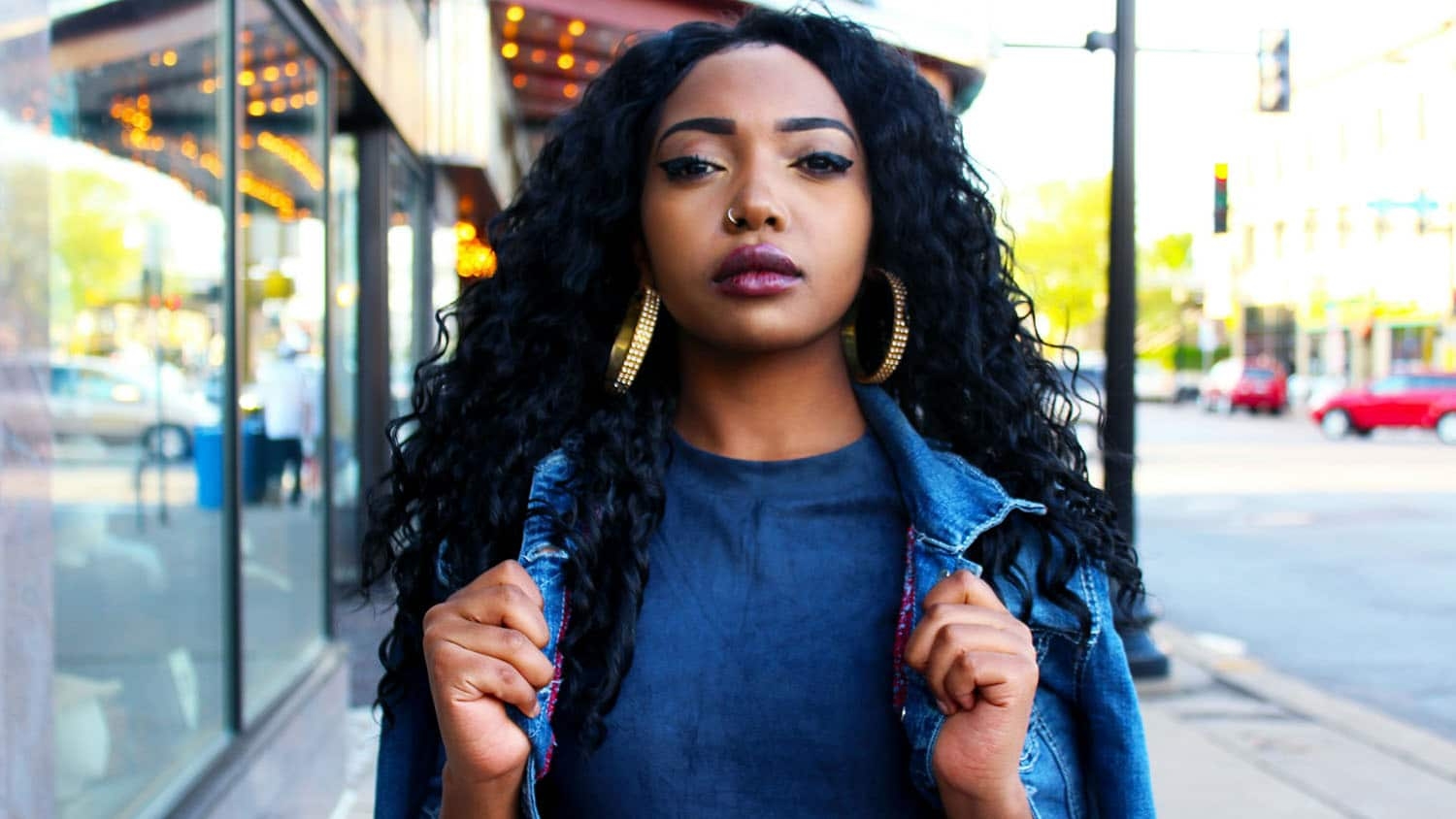 a young Black woman looks directly at the viewer while standing on a city street